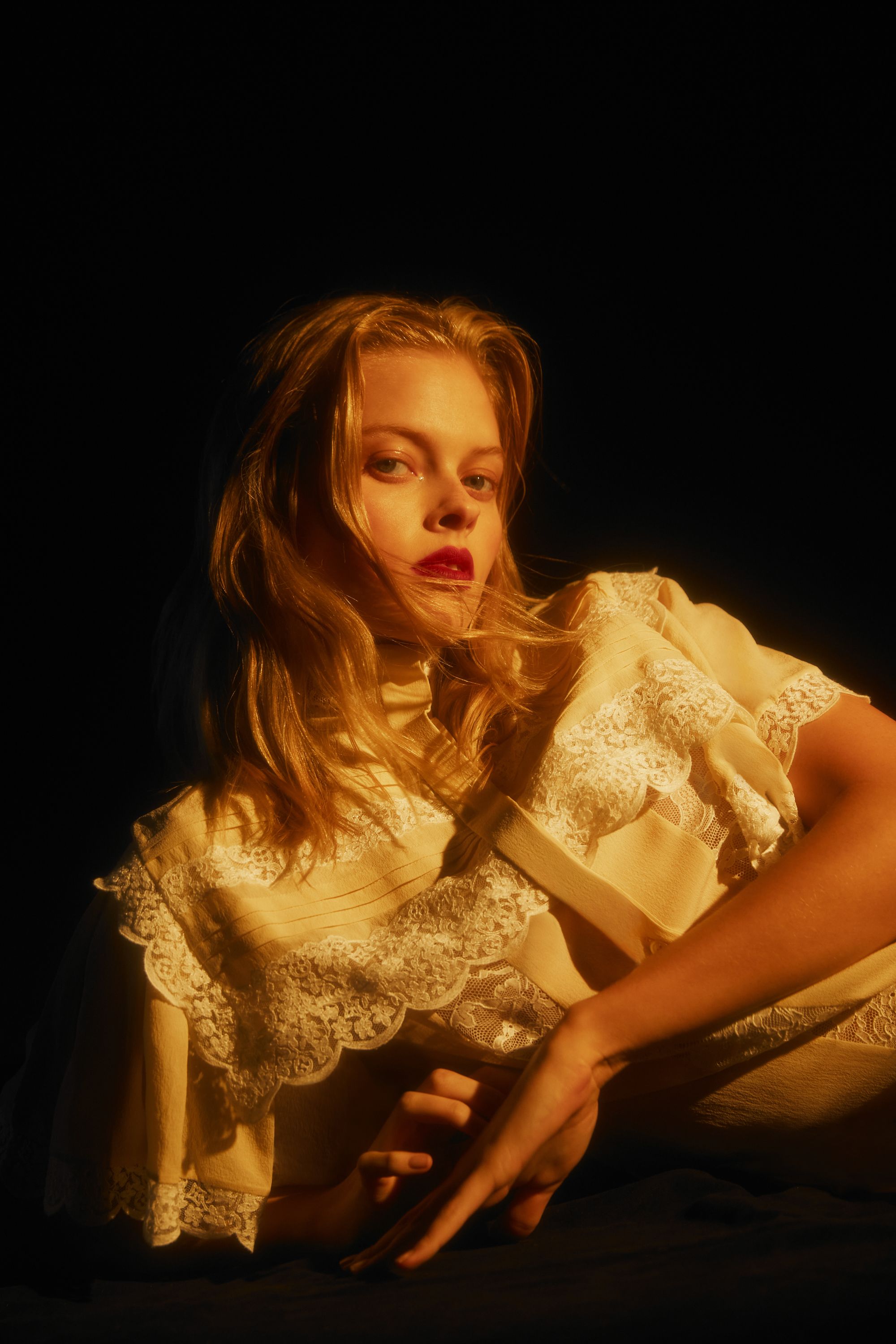 Blonde woman with red lipstick poses in a cream-colored lace dress against a dark background with moody golden lighting, creating a dramatic fashion image.