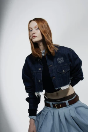 Red-haired model wearing a dark denim jacket and light blue skirt with a brown belt against a white studio background.