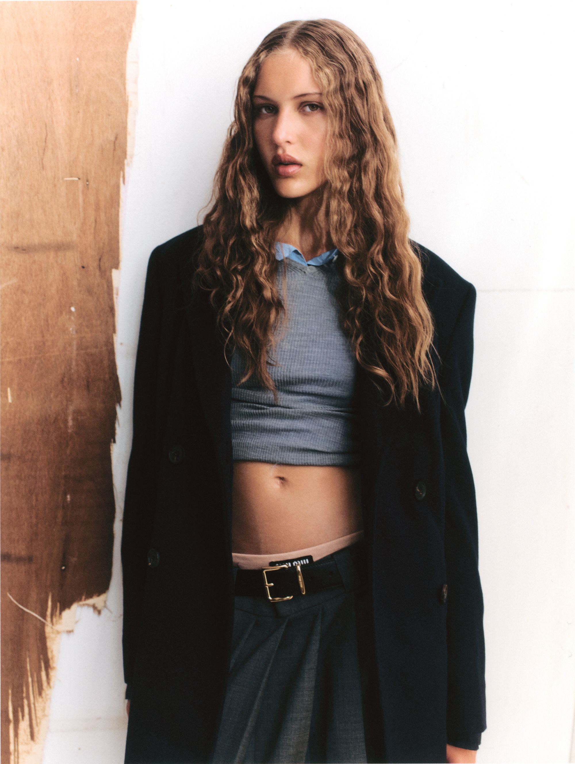 Young woman with long wavy hair posing in an indoor environment wearing a gray shirt, dark skirt, and oversized black blazer.