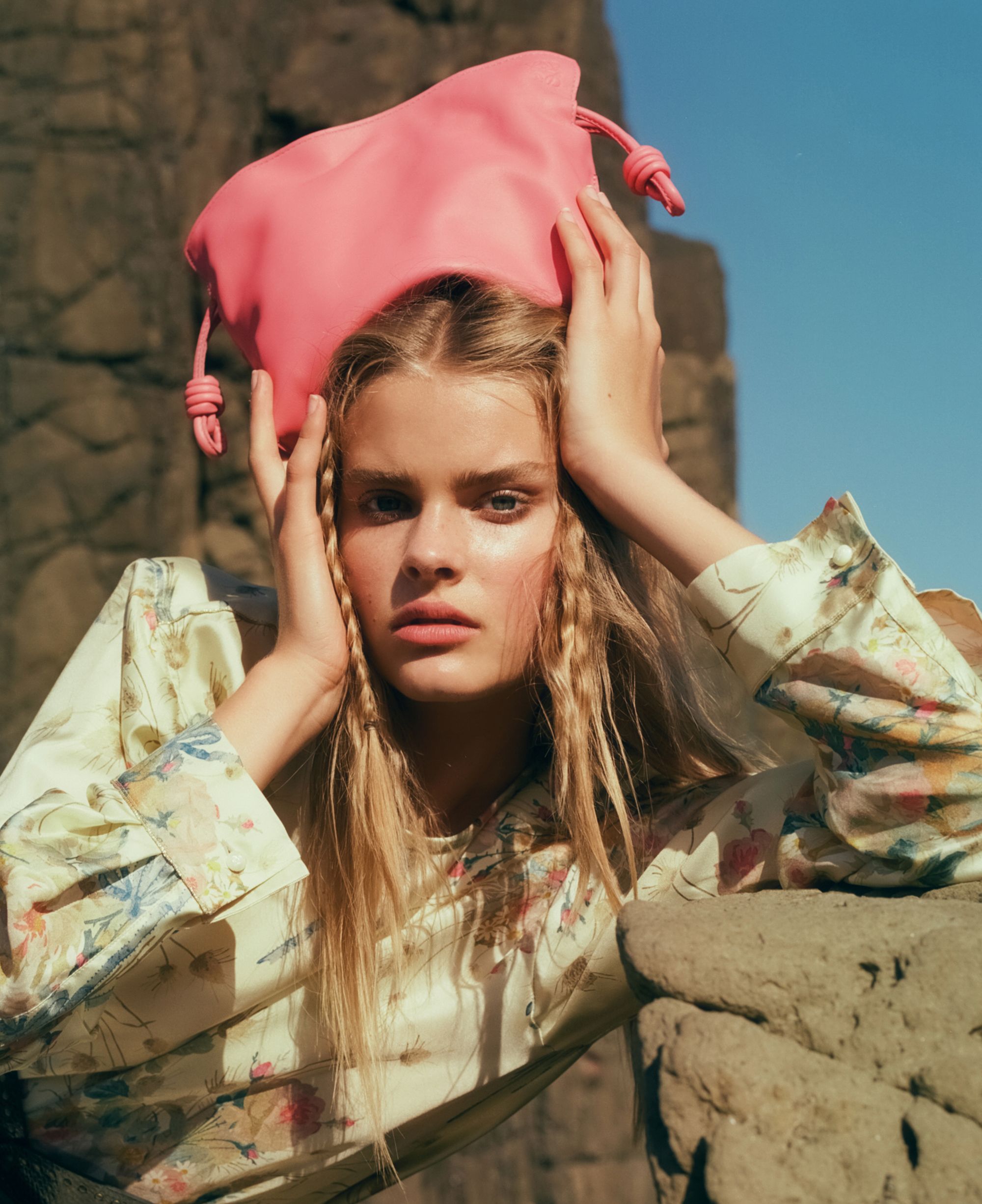 Close-up of a woman with light skin and crimped blonde hair, posed outdoors holding a pink handbag above her head.