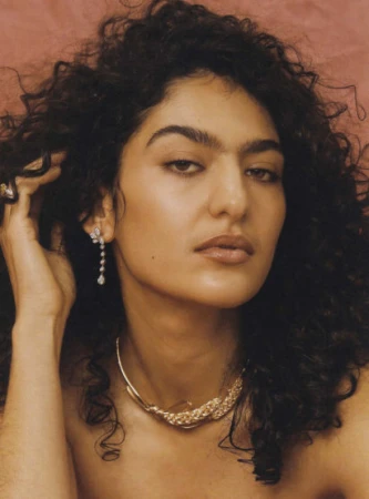 Close-up portrait of a woman with dark curly hair wearing a gold choker necklace and diamond drop earrings against a warm background.