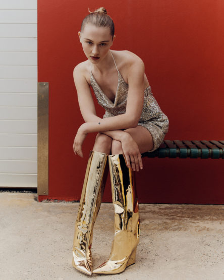 Adult female sitting on a bench in a sparkly mini-dress and gold, knee-high boots in front of a red and white paneled wall.