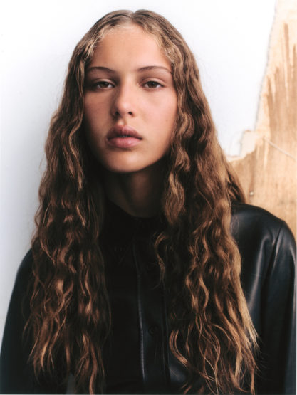 Portrait of a young woman with long wavy brown hair wearing a black top, set against a white and brown textured background.
