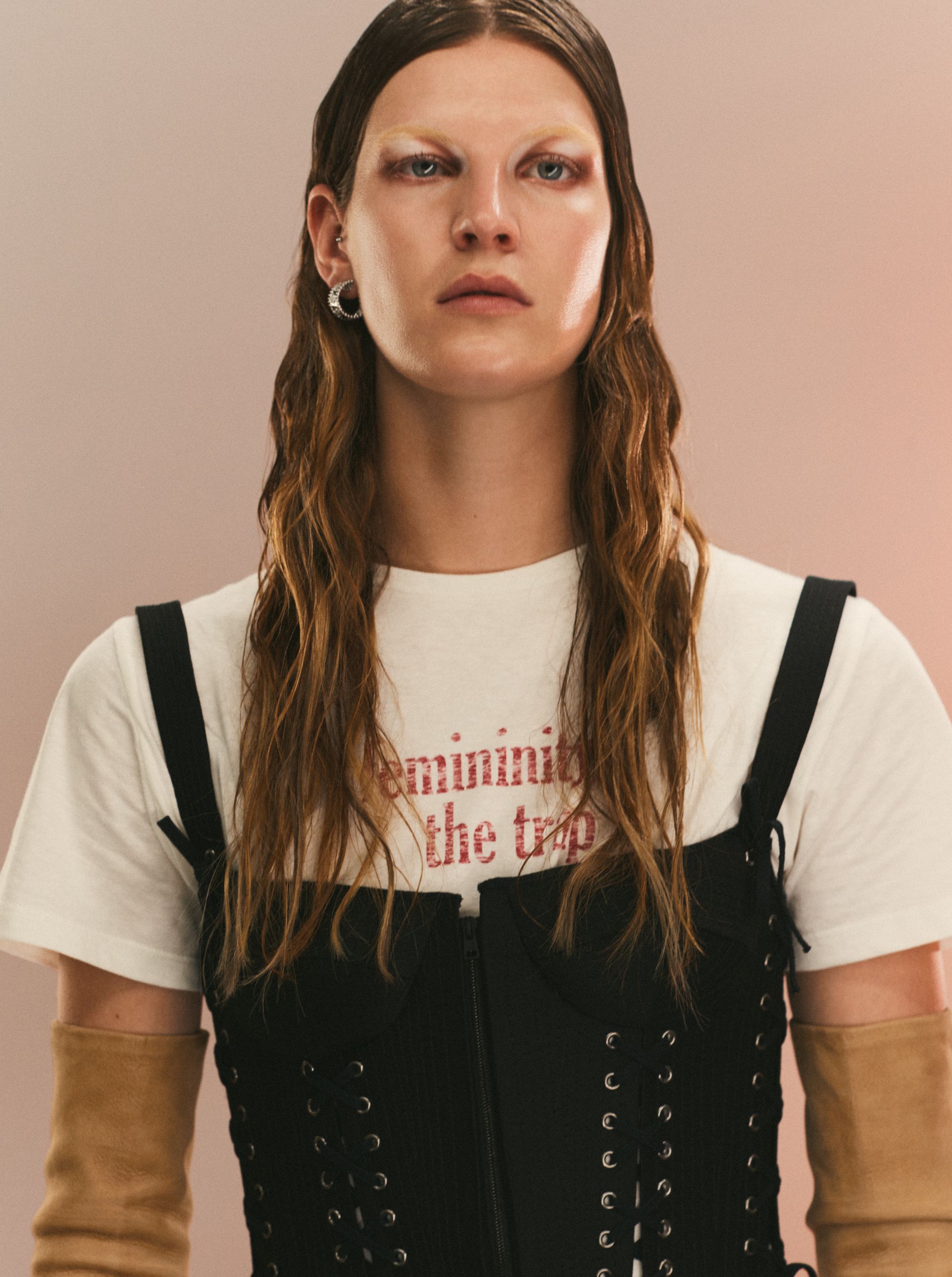 Portrait of a fair-skinned adult with long, wavy brown hair, wearing a white T-shirt and black corset against a gradient pink background.