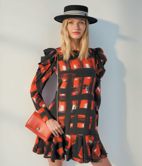 A fashionable adult woman in a red and black patterned dress stands in a studio holding a red clutch bag and wearing a black hat.