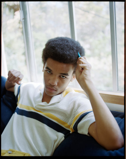 Indoor portrait of a young adult Black male with an afro hairstyle, wearing a striped shirt, seated on a blue cushion by a window.