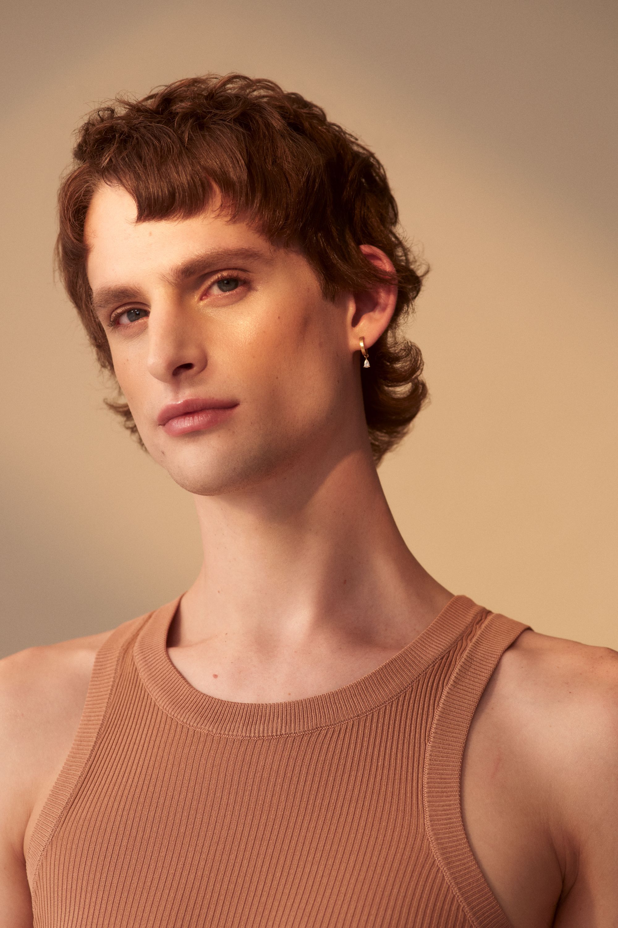 Close-up portrait of an adult male with brown hair and a tan ribbed tank top in front of a neutral background with soft lighting.