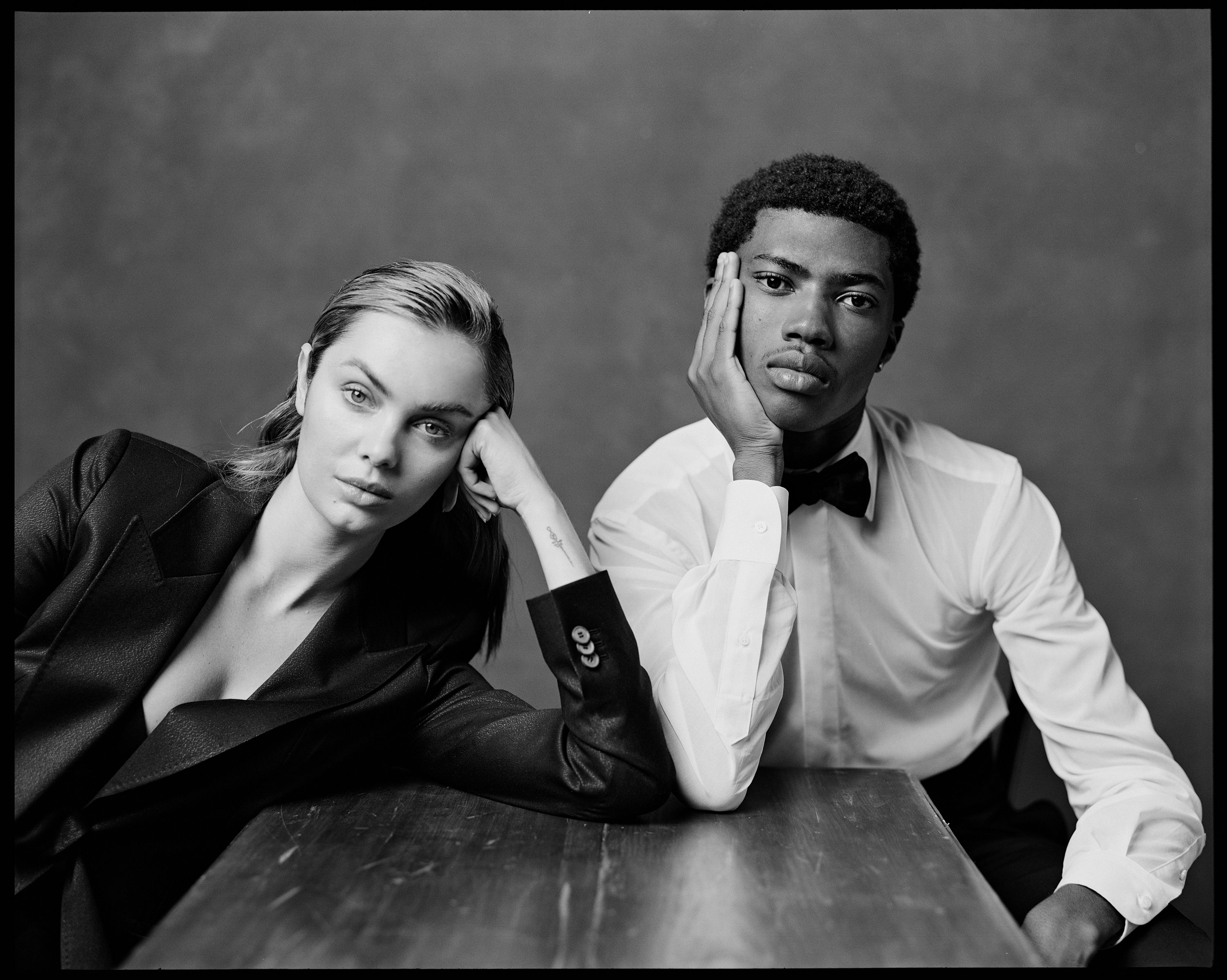 Black and white studio portrait of a Caucasian female in a blazer and an African-American male in formalwear at a wooden table.