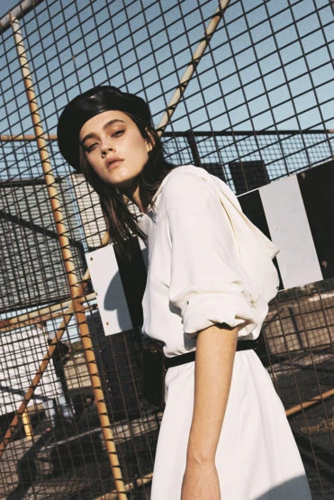 A young woman in a white dress and black beret poses against a metal fence with a partly cloudy sky in an outdoor setting.