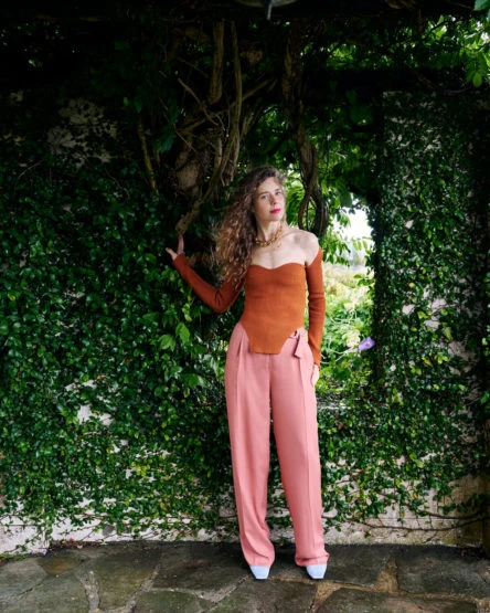 Adult woman in a copper-colored top and rose-colored pants stands against a textured stone wall covered in green vines and greenery.