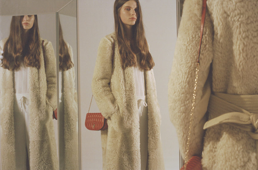 Indoor fashion portrait featuring an adult woman wearing a cream-colored coat and carrying a red purse, reflected in a nearby mirror.