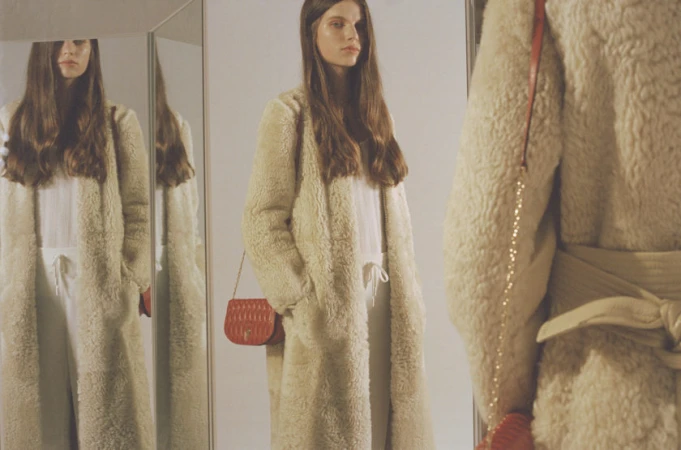 Model with long brown hair wearing a cream shearling coat and holding a red crossbody bag, reflected in multiple mirrors.