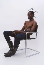 Shirtless black man with sculpted braided hair sitting on a chrome chair wearing dark jeans and cowboy boots against a white wall.