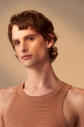Portrait of a man in his 20s with short textured brown hair wearing a beige ribbed tank top against a warm gradient background.