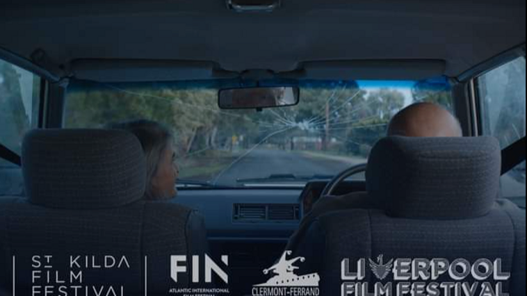 Inside a car, an older couple drives down a tree-lined road with a cracked windshield, captured from the back seat perspective.
