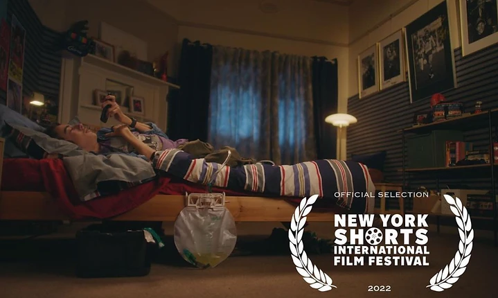 Teenager lying on a bed using a smartphone in a dimly lit bedroom with striped walls and curtains, plus the New York Shorts film festival logo.