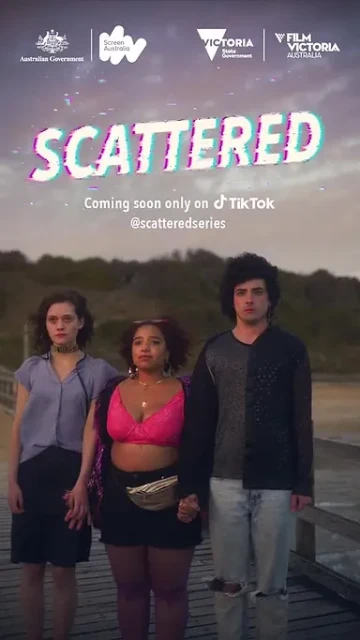 Three young adults stand holding hands on a boardwalk near a beach with trees in the background in a promotional still for 'Scattered'.