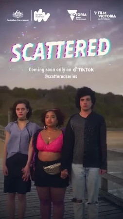Three young adults stand holding hands on a boardwalk near a beach with trees in the background in a promotional still for 'Scattered'.