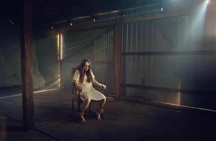 Woman in a vintage white dress bound to a chair in a rustic, shadowy interior with light streaming through metal siding.