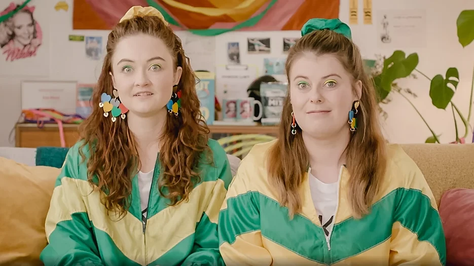 Two women in green and yellow jackets, seated indoors, smile toward the camera wearing colorful earrings and green hair scrunchies.