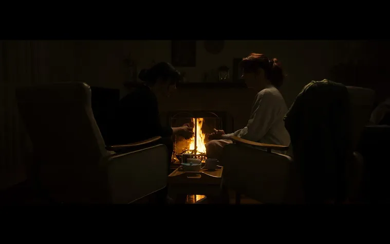 Two women in armchairs sit near a glowing fireplace, their hands outstretched, illuminated by the warm light in a dark room.