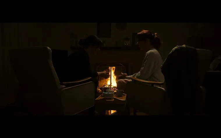 Two women in armchairs sit near a glowing fireplace, their hands outstretched, illuminated by the warm light in a dark room.