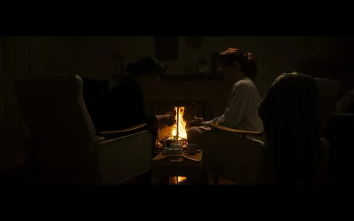 Two women in armchairs sit near a glowing fireplace, their hands outstretched, illuminated by the warm light in a dark room.