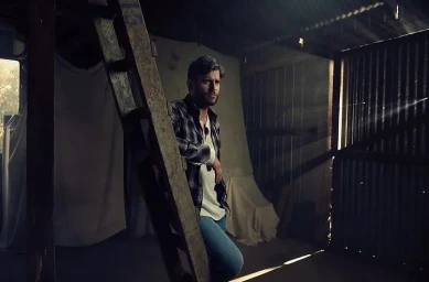 A man in a plaid shirt and blue jeans leans against a wooden ladder inside a rustic barn with corrugated metal walls.