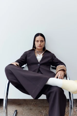 Model with dark hair seated in a chrome tubular frame chair wearing a dark brown blazer and trousers against a white wall.