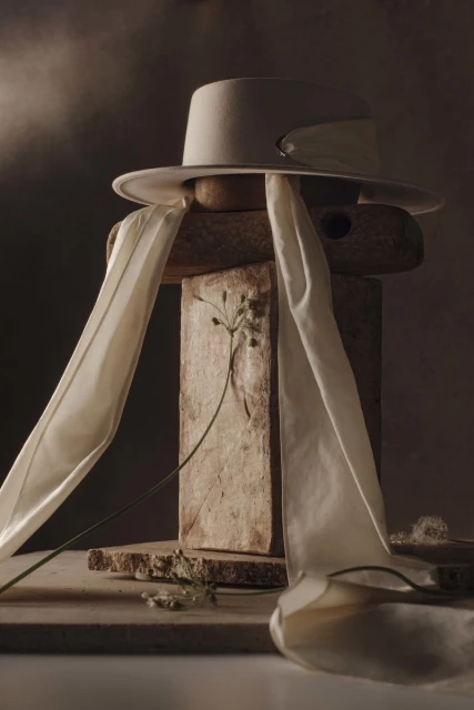 Beige hat with white silk scarf on a stack of raw stone blocks, next to a sprig of wild grass against a neutral brown background.