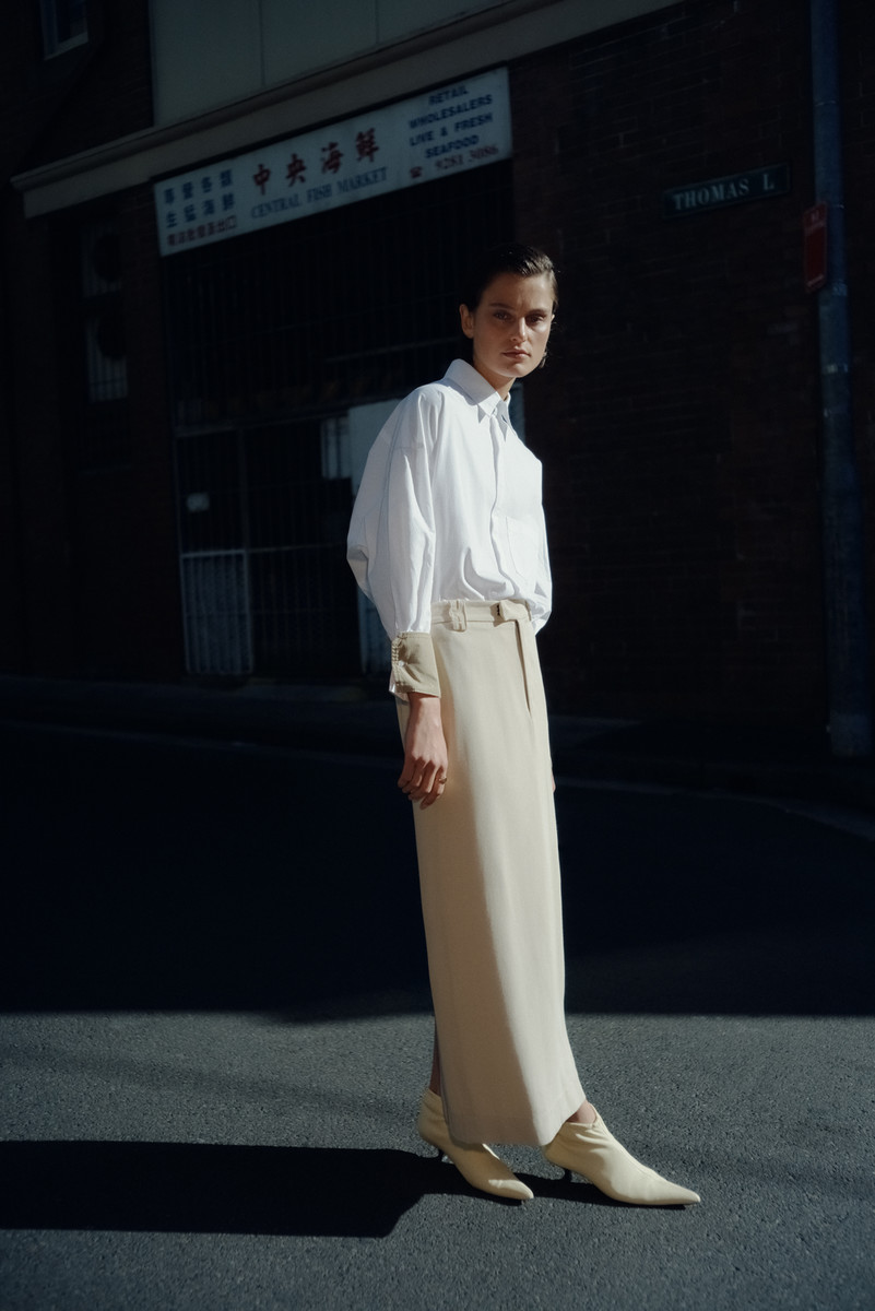 A woman poses in a white shirt and tan skirt on a city street with an Asian market, bathed in sunlight and showcasing fashion-editorial style.