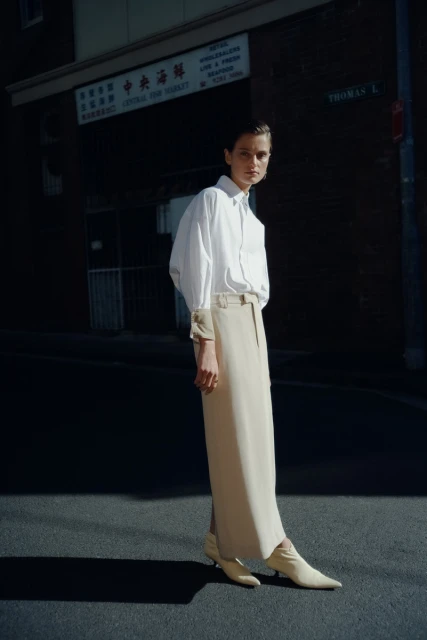 A woman poses in a white shirt and tan skirt on a city street with an Asian market, bathed in sunlight and showcasing fashion-editorial style.