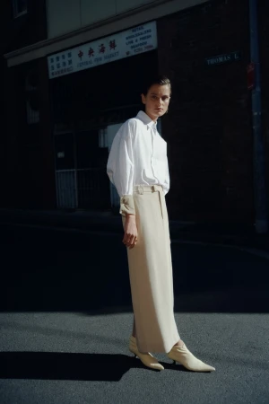 A woman poses in a white shirt and tan skirt on a city street with an Asian market, bathed in sunlight and showcasing fashion-editorial style.