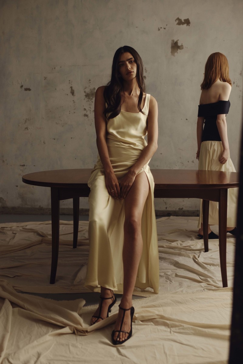 Two women in a fashion shot are posing in front of a table and grey wall wearing flowing dresses and heels with soft lighting.
