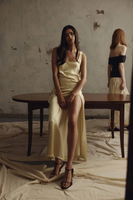 Two women in a fashion shot are posing in front of a table and grey wall wearing flowing dresses and heels with soft lighting.