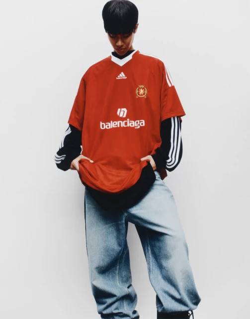 Young person in red Balenciaga and Adidas branded t-shirt and denim baggy jeans standing against a white wall in a fashion editorial.