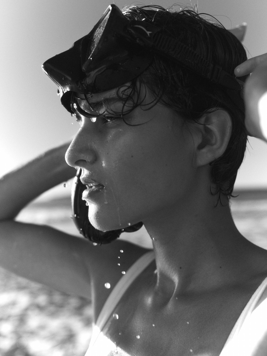 Black and white portrait of a wet woman wearing a diving mask, set against a blurred ocean and sky background, captured in natural light.