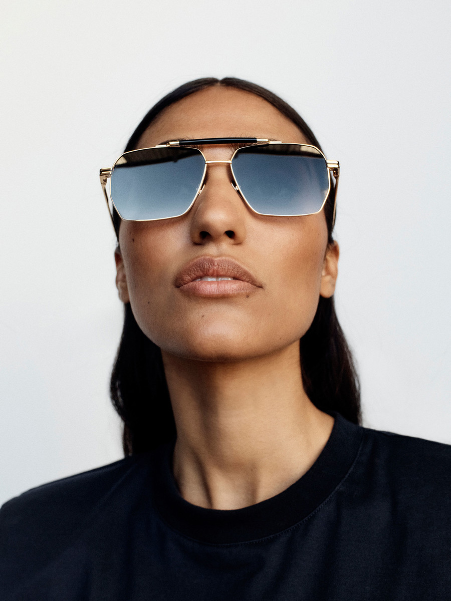 Portrait of a woman with dark hair and tanned skin wearing stylish gold sunglasses against a plain white backdrop in a studio setting.