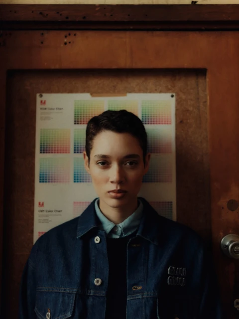 Woman with short hair wearing a denim jacket posing indoors against a color chart and a brown painted doorframe in a studio.