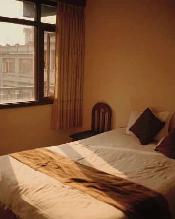 A bedroom interior with a made bed, wooden chair, window with curtains, and warm morning light casting long shadows.