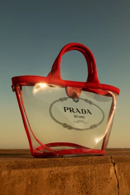 Clear tote bag with red trim and Prada logo on a concrete surface against a gradient blue and yellow sky background.