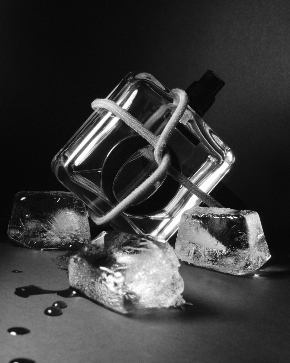 Black and white studio shot of a perfume bottle wrapped with white string surrounded by three ice cubes and water droplets.
