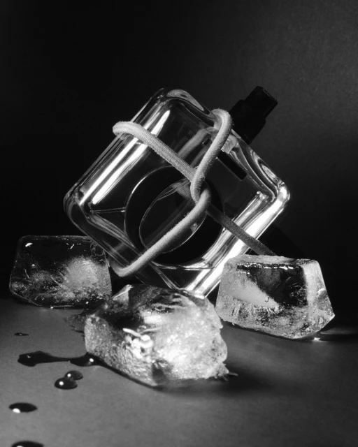 Black and white studio shot of a perfume bottle wrapped with white string surrounded by three ice cubes and water droplets.
