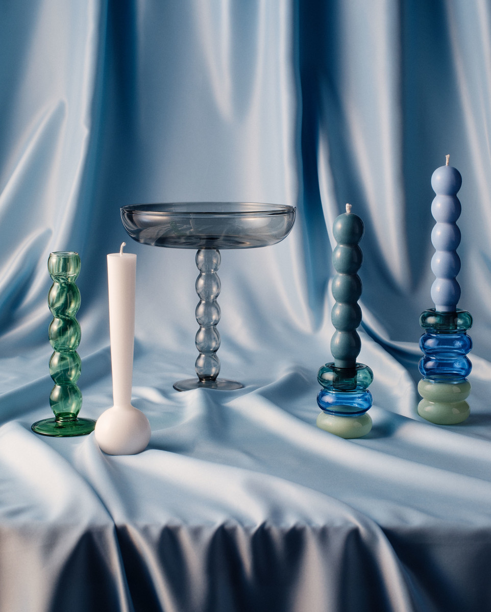 Still life of five candles and decorative glass stands of different shapes and sizes, presented on a draped blue fabric.