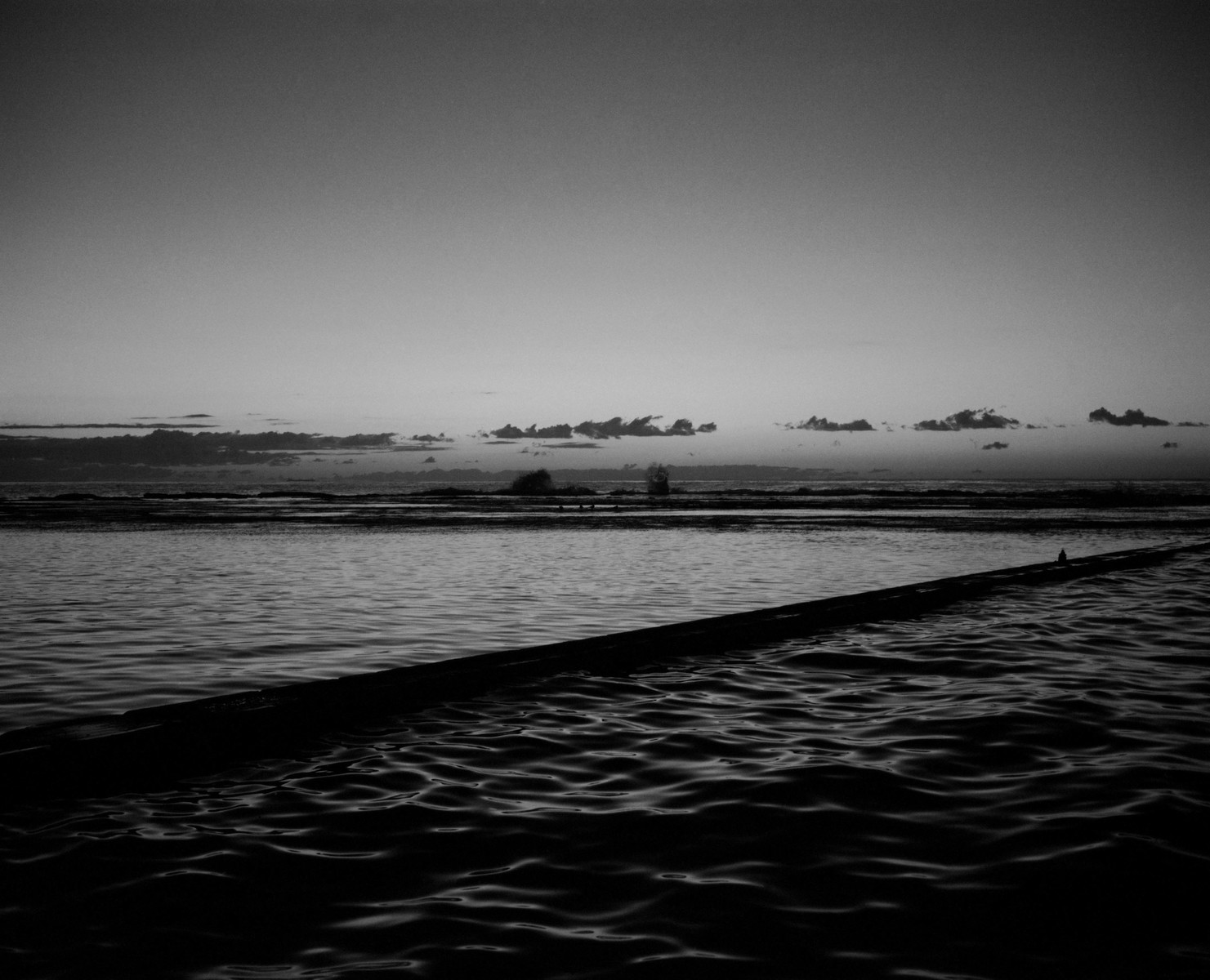 Monochrome seascape featuring a still body of water under a cloudy sky, with a wooden structure extending into the water.