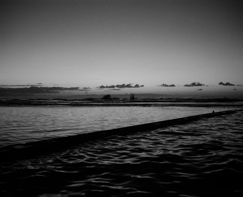 Monochrome seascape featuring a still body of water under a cloudy sky, with a wooden structure extending into the water.