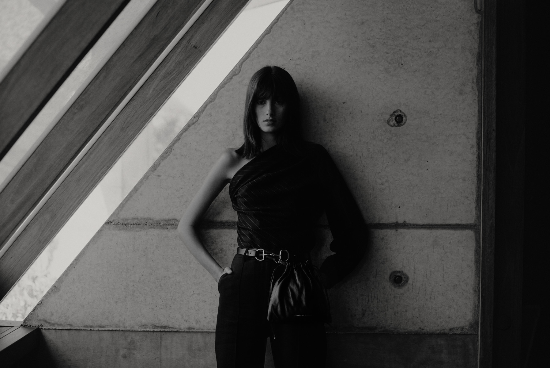 Monochrome fashion shot of a young woman in one-shoulder top and high-waisted pants leaning against a concrete wall with window frame.
