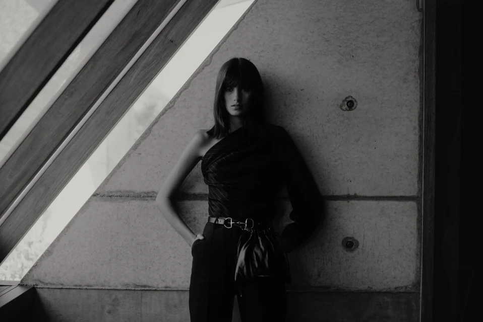 Monochrome fashion shot of a young woman in one-shoulder top and high-waisted pants leaning against a concrete wall with window frame.