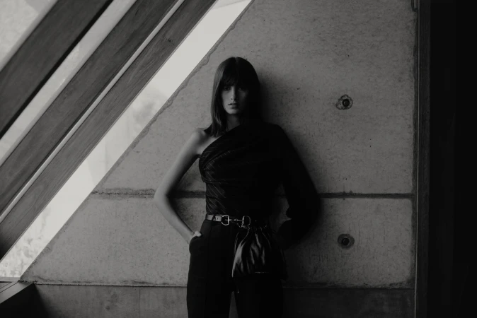 Monochrome fashion shot of a young woman in one-shoulder top and high-waisted pants leaning against a concrete wall with window frame.