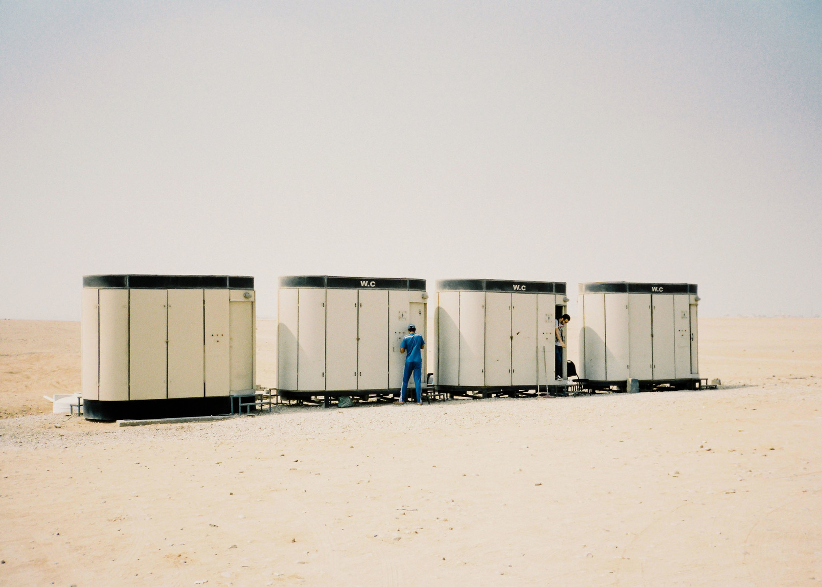 Four portable restrooms stand in the desert landscape, each with a 'WC' sign, and two men are interacting with the facilities.